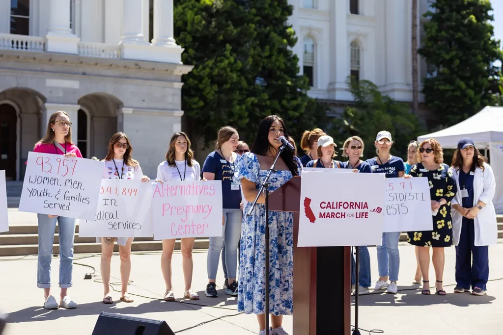 A speaker at the California March for Life podium outside the State Capitol, with pregnancy center supporters holding signs of their results.